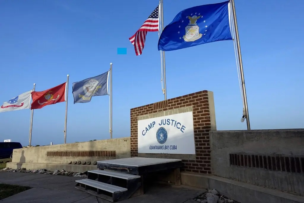 Flags for the U.S. armed forces and the American flag fly above a brick sign that says Camp Justice, Guantnamo Bay, Cuba.