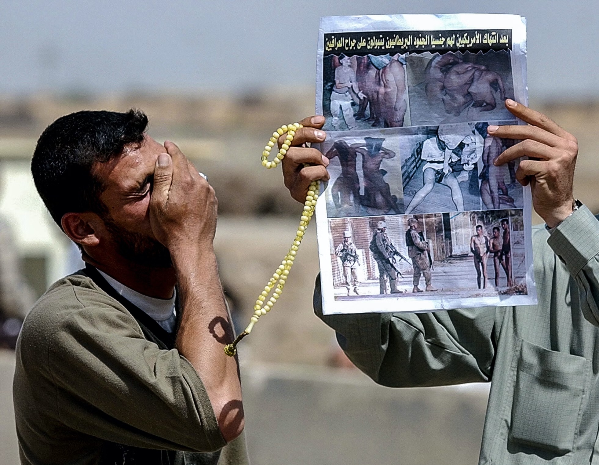 A relative of an Iraqi prisoner being held by US authorities at the Abu Ghraib prison holds his hand to his face 08 May 2004 as another one shows a newspaper featuring photos of US soldiers abusing Iraqi prisoners inside the detention center located 30 kms west of Baghdad.  Hundreds of Iraqis gather in front of the jail daily in the hope of getting news about their loved ones.    AFP PHOTO/Roberto SCHMIDT / AFP / ROBERTO SCHMIDT        (Photo credit should read ROBERTO SCHMIDT/AFP via Getty Images)