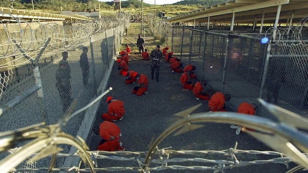 Detenidos vestidos con uniformes naranjas, están esposados y agachados en el suelo de un recinto exterior cerrado por altas vallas con alambre de espino. Hay varios soldados que los vigilan.