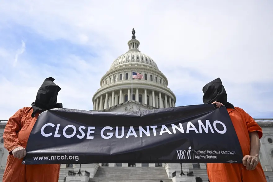 people dressed in orange jumpsuits with bags over heads hold sign saying close guantanamo in front of the US capitol